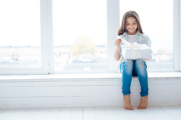 Great present. Window sill becomes recreating place for happy attractive little girl  who unpacking her present while looking at it