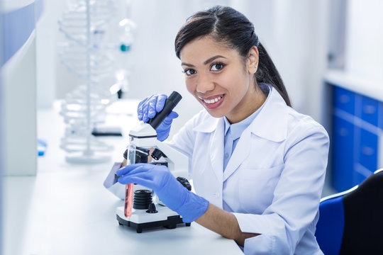 Biological studies. Delighted positive nice woman holding a microscope and smiling while doing biological research