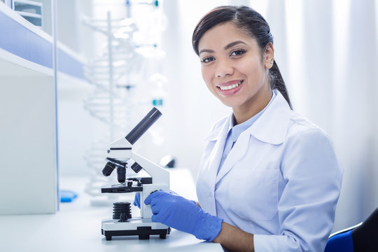 Scientific equipment. Delighted smart nice woman sitting in front of the microscope and looking at you while doing a biological research