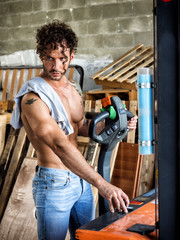 Young shirtless man with muscular body in workshop using forklift. 