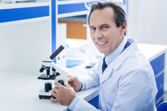 Biological research. Positive nice experienced man sitting in front of the microscope and looking at you while working in the lab