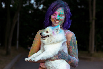 Merry brunette woman holding little white dog covered colorful Holi powder