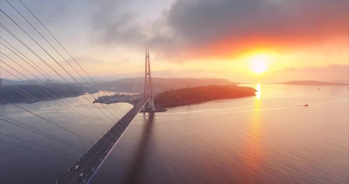 Flying backwards, aerial view of Russian island and cable-stayed Russian Bridge across the Eastern Bosphorus strait in Vladivostok, Russia. Amazing bright sunrise