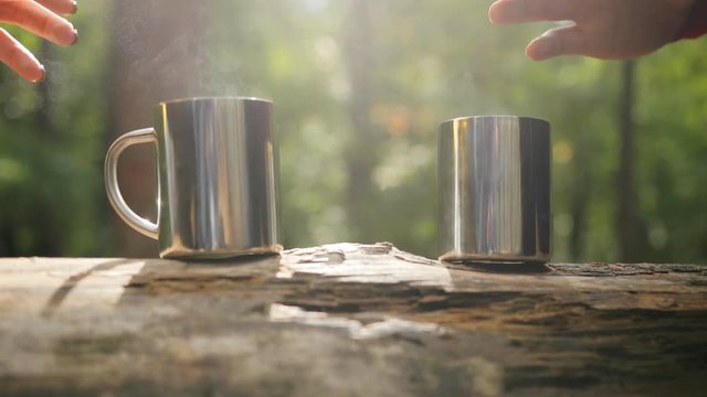 Close up of the tw cups with a hot tea on the trunk being taken by male and female hands. Forest on the background. Outdoor