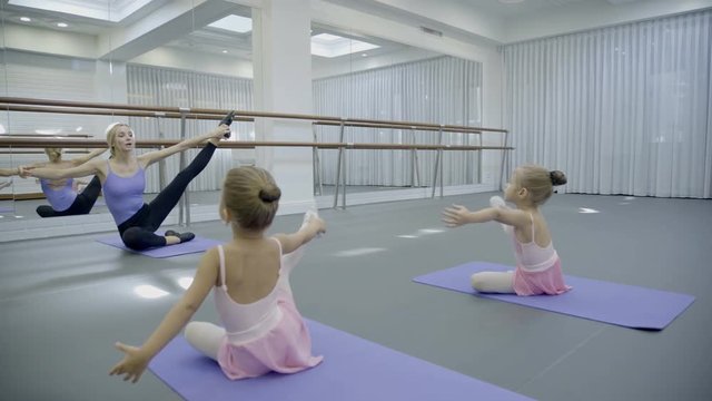The blond ballet teacher leads the stratching with her two little students in the bright gym. Girls and ballerina sit on the mats with left leg put under the body and taking the right one up sideways