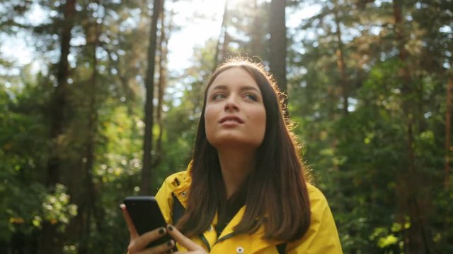 Portrait shot of young beautiful woman strolling in the forest and stopping to take photos of the nature on the black smartphone. Sunny weather. Outdoor