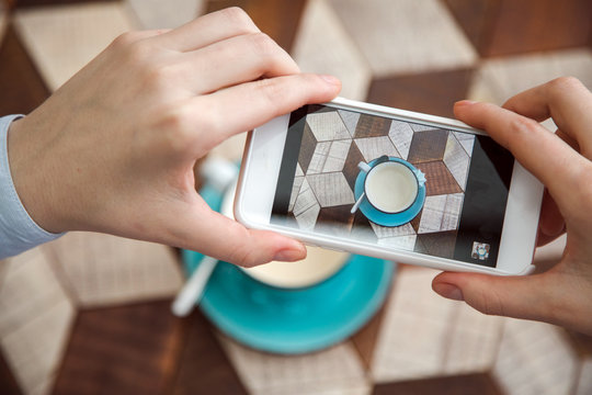 Blogger takes pictures of coffee on table indoor. Close-up hand holding phone mobile taking photo cappuccino