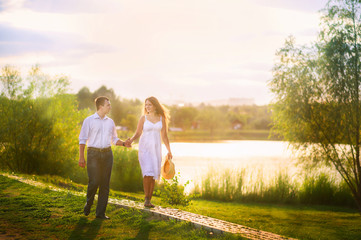 Beautiful summer picture on the nature by the river. A loving couple runs along the grass against the backdrop of a pond. A girl in a white sarafan and a straw hat and a guy in a white shirt.