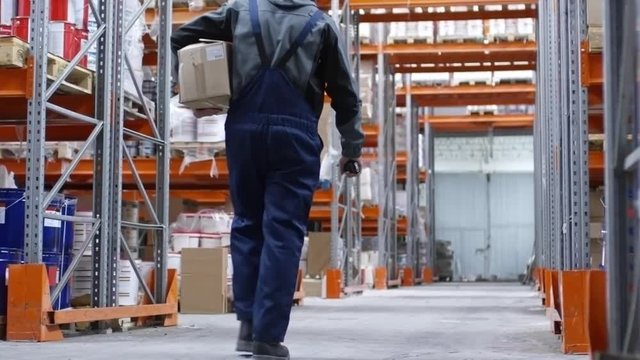 PAN of male worker in hard hat carrying cardboard box and walkie-talkie walking through aisle of warehouse with rack shelves