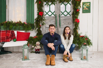 Obraz na płótnie Canvas Happy beautiful young couple in love dressed in sweater sitting on porch steps at light house with decorated in red green New Year door at home. Christmas or birthday pipes. Family and holiday 2018