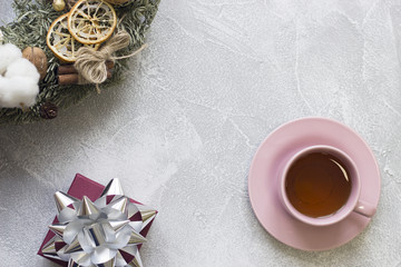 Pink tea cup, saucer, purple gift box with a silver shiny bow, christmas pine wreath on a light concrete background, top view