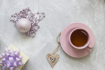 Obraz na płótnie Canvas Pink tea cupwith a saucer,a white gift box with a purple bow, pink shiny ball, pink star-shaped beads and a gingerbread on a light concrete background, top view