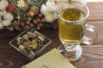 A glass tea cup with a green tea ,a square bowl of dry flowers, a Christmas wreath and a gift box on a wooden background
