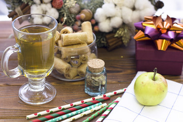 A glass cup of green tea, a bowl of cookies, a tiny jar with dry flowers, a purple giftbox, a green apple on a white plaid napkin, patterned straws and a Christmas wreath on a wooden table