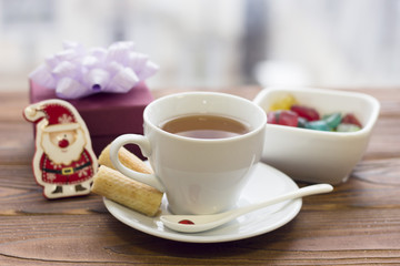Black tea in a white porcelian cup with saucer, white teaspoon, a bowl of colored candies, a purple gift box with a light purple bow and a toy santa on a wooden table