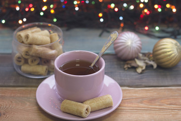 Festive still life with a cup of tea, bowl of cookies, pink and golden Christmas-tree balls and fairy lights, colored wooden background
