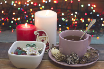Festive still life with a pink porcelian cup of tea, a saucer, a teaspoon, bowl of colored candies,two burning candles and fairylights on the back, a colored wooden background