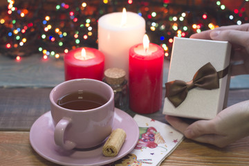 Hand holding a white gift box with a brown bow, a pink porcelian cup of tea, a saucer, two red and one white burning candles and fairylights on the back, a colored wooden background