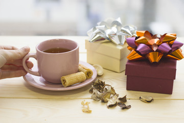 A hand holding a pink cup of hot black tea,a purple gift box with a shiny bow, a light giftbox with a silver bow, dry flowers on a wooden table, light background