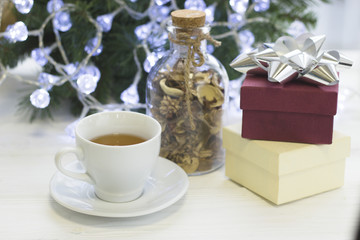 A festive bright morning with a white porcelian cup of hot black tea, a glass clear bottle of dry flowers, a light gift box and a purple gift box with a silver bow, Christmas tree with white fairy
