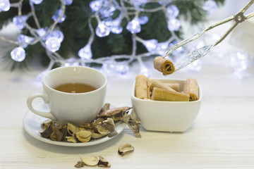 Hand holding a cookie with a tong above a bowl of cookies, a white porcelian cup of hot black tea, dry flowers spilled on a saucer,a blurred Christmas tree with white fairy lights on the back, light