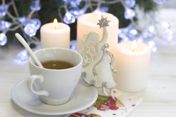 Obraz na płótnie Canvas A festive still life with a white tea cup, a spoon and a saucer, a white porcelian angel, tree white burning candles, a blurred Christmas tree with fairy lights on the back, light background