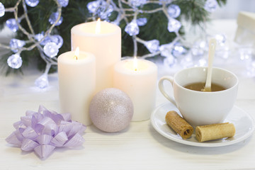 Fototapeta na wymiar A festive still life with a white tea cup, a spoon and a saucer, a shiny pink Christmas-tree ball, a purple bow, tree white burning candles, a blurred Christmas tree with fairy lights on the back