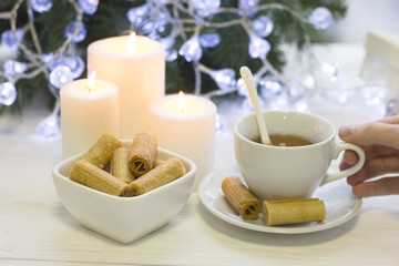 Hand holding a wite cup of tea, a calming flame of three white burning candles, a white bowl of fresh cookies, blurred Cristmas tree with fairy lights on the back, light background