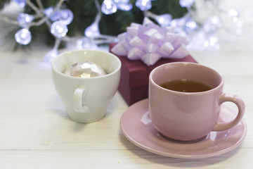 A festive still life with pink porcelian tea cup with a sacer, a dark purple giftbox with a violet bow and Christmas-tree ball in a white tea mug, blurred Christmas-tree with fairy lights on the back