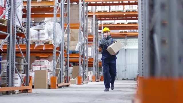PAN of male worker in hard hat carrying cardboard box and talking via two-way radio with colleague while walking through warehouse aisle with rack shelves