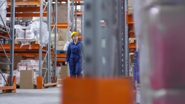 PAN of concentrated female worker in glasses and hard hat walking throug warehouse aisle, then stopping and writing down something on clipboard while checking inventory