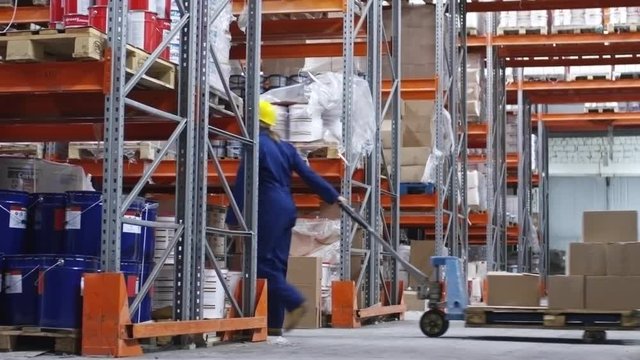 PAN of female worker wearing hard hat pulling cart with cardboard boxes and walking through factory warehouse with rack shelves