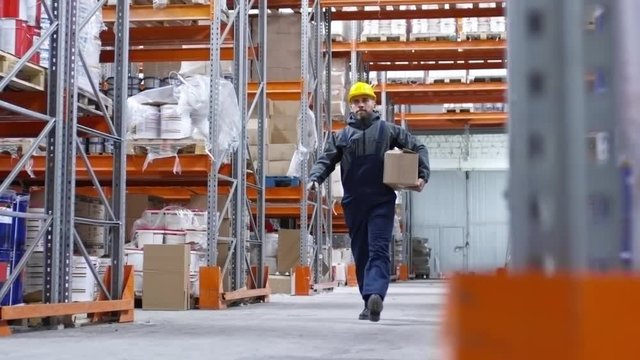PAN of bearded male worker in hard hat carrying walkie-talkie and cardboard box and walking through aisle with rack shelves in factory warehouse