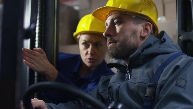 Tilt up of female warehouse manager teaching male worker in hard hat how to operate forklift