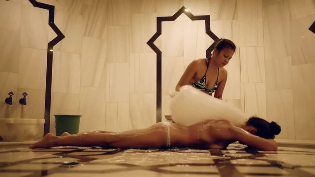 naked tanned brunette is lying on a stone table in hammam and masseur is covering her body by foam