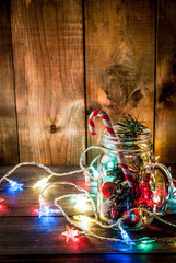 Christmas, New Year's concept. Mason Jar with Christmas decorations, fir cones, artificial snow, candy cane and fir branch. On a wooden table background, with a lit garland turned on. Copy space