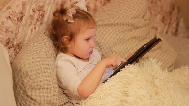 Funny child looks at the screen and plays downloaded application on tablet laptop, computer A little cute girl lies in bed in a bedroom under the blanket, looking cartoon and playing the game.