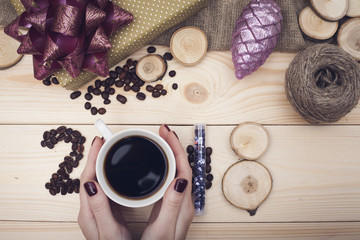 2018 inscription of coffee beans, woman's hands holding a coffee mug and wooden slices, purple christmas toys with a skein of threads on a light wooden background, top view