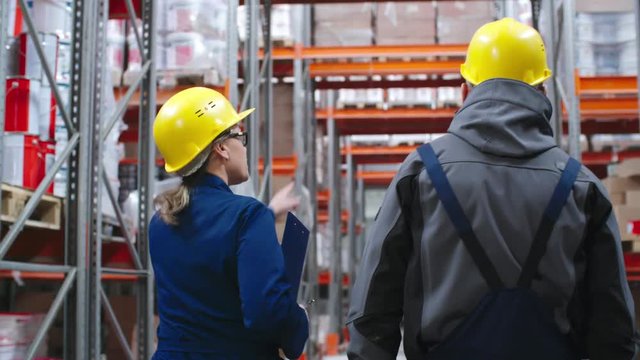 Tracking with rear view of male worker in hard hat holding walkie-talkie and talking with serious female colleague in uniform while walking through factory warehouse