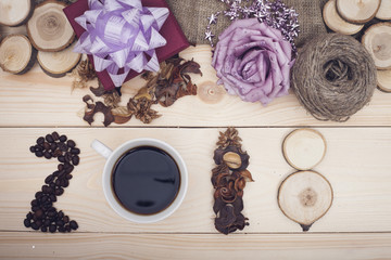 Fototapeta na wymiar 2018 inscription of coffee beans, cup, dry flowers and wooden slices, a dark purple gift box with a violet bow, purple beads and a rose, a skein of threads, light wooden background, top view