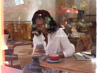 Attractive natural beauty african american woman with long curly hair drinks coffee in modern hipster cafe, works on laptop and makes selfie or sends texts on smartphone uses internet technology