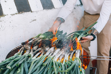 Man in white tshirt and with authentic arm tattoos, makes barbecue on grill on outside terrace of spring onions, calcots or cambray as traditional spanish, catalan or mexican food - obrazy, fototapety, plakaty