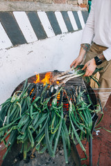Obraz premium Man in white tshirt and with authentic arm tattoos, makes barbecue on grill on outside terrace of spring onions, calcots or cambray as traditional spanish, catalan or mexican food