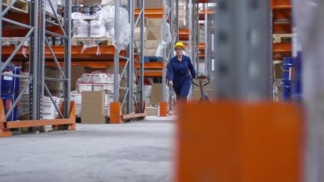 PAN of concentrated female worker wearing glasses and hard hat pulling cart with cardboard boxes and walking between shelves in factory warehouse