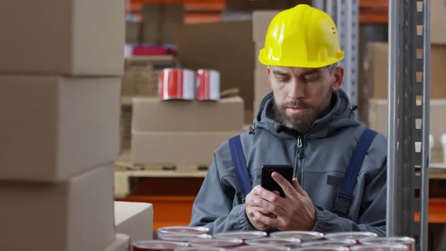 Zoom out with slowmo of bearded male warehouse worker in hard hat stocking cans of paint and calling something on mobile phone