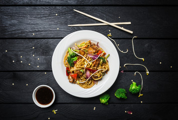 Wok. Udon stir fry noodles with chicken and vegetables in a white plate on black wooden background. With chopsticks and sauce