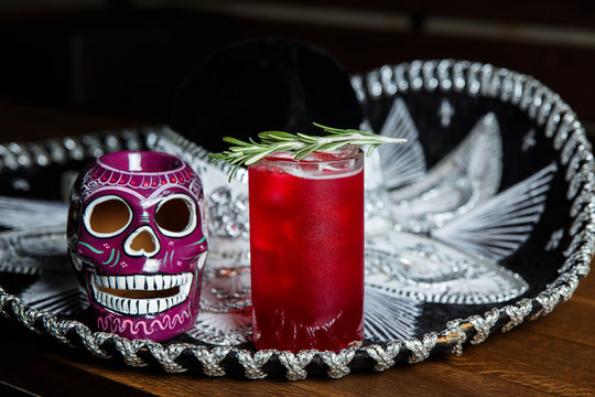 A horizontal image of a red cocktail with ice cubes in a highball glass, decorated with rosemary. Sombrero and a mexican colourful skull near by.