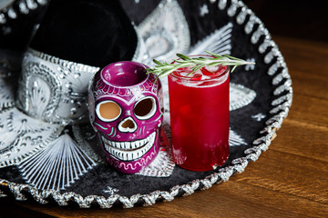 A horizontal image of a red cocktail with ice cubes in a highball glass, decorated with rosemary. Sombrero and a mexican colourful skull near by.