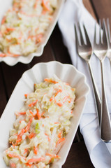 Freshly homemade, traditional American cole slaw made of cabbage, carrots, mayonnaise, vinegar, sugar, salt and pepper in white bowl on wooden table next to white cloth napkin and some silver forks