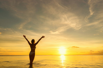 Back of young woman standing in water with raised arms and looking at sea and sunset.